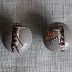 Puede incluir: Dos pomos decorativos redondos y grises con ilustraciones de animales pintadas a mano. Uno presenta una criatura parecida a un zorro, el otro un animal parecido a una jirafa sosteniendo flores. Los pomos son probablemente para muebles o decoración.