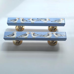 May include: Two blue wooden cabinet pulls with painted seagulls. Each pull has a light blue background with five white and gray seagulls. The pulls are mounted on gold-colored metal bases.