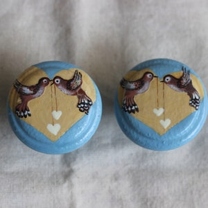 Könnte beinhalten: Zwei blaue und gelbe herzförmige Knöpfe mit gemalten Vögeln und weißen Herzen. Die Vögel schauen sich an und scheinen eine Schnur mit daran hängenden Herzen zu halten.