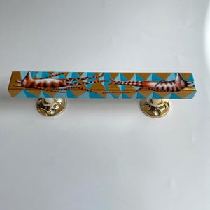 Pode incluir: Um puxador decorativo para armário com uma barra retangular de madeira e uma ilustração de gato. A barra tem um fundo dourado com formas de diamante azul. O gato é marrom e branco. O puxador tem dois botões dourados.
