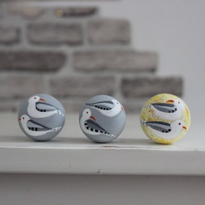 May include: Three decorative wooden spheres with a painted seagull design. Two spheres are light gray, and one is yellow with a speckled pattern. Each sphere features two white seagulls with gray wings and red beaks.