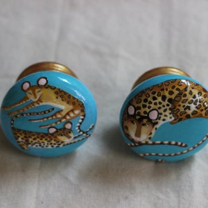 May include: Two round, blue, ceramic knobs with gold accents. One knob features a painted design of two leopards, one on top of the other. The other knob features a single leopard painted in a curled position.
