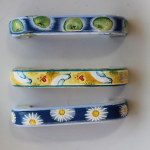 May include: Three decorative drawer pulls with painted designs. The top pull features green apples on a blue background. The middle pull has a yellow background with blue birds and red hearts. The bottom pull has a blue background with white daisies.