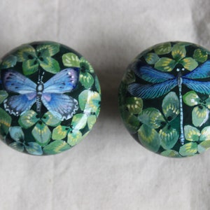 May include: Two round, green painted ornaments with a clover leaf design. One ornament features a blue butterfly and the other features a blue dragonfly.