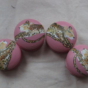 May include: Four pink wooden knobs with hand-painted images of leopards. Each knob features a different pose of the leopard, with intricate details in brown and black.