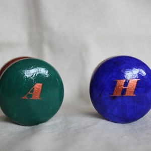 May include: Two decorative wooden spheres are shown, one green with a copper-coloured "A", the other blue with a copper-coloured "H". A third sphere is visible in the background. These spheres are likely decorative accents.