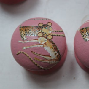 May include: Two pink round knobs featuring a design of leopards and a smaller spotted animal. The knobs are painted with a pink background and the animals are painted in brown, black, and white.