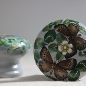 K&ouml;nnte beinhalten: Ein Satz von drei Keramik-Schubladengriffen mit einer hellgrauen Basis und handbemalten Schmetterlings- und Blumenmotiven. Die Schmetterlinge sind braun mit schwarzen Markierungen und die Blumen sind wei&szlig; mit gelben Zentren.