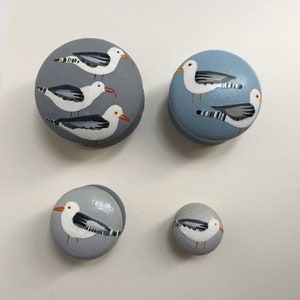 Peut inclure: Quatre ornements ronds en bois peints avec des mouettes. Les ornements sont dans des tons de gris et de bleu. Trois des ornements ont trois mouettes chacun, tandis que le plus petit ornement a une mouette.