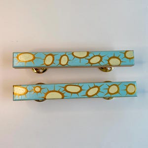 Peut inclure: Deux barrettes rectangulaires bleu clair avec un motif répété de formes dorées en forme de soleil. Chaque barrette a un fermoir en métal doré au dos.