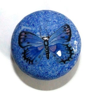 May include: A round, blue decorative stone featuring a painted butterfly design. The butterfly is rendered in shades of blue, black, and purple, set against a textured blue background. This is a decorative item.