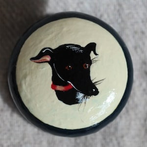 May include: A round, hand-painted decorative knob featuring a black dog with a red collar on a cream-colored background. The knob has a dark blue border and is likely made of wood or ceramic.