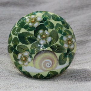 May include: A round decorative knob with a pale yellow base, decorated with green leaves, white flowers, and a snail. The knob is likely ceramic or similar, suitable for furniture or cabinet doors. The diameter is approximately 4 cm.