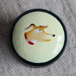 May include: A round, painted object featuring a dog's head. The dog has tan fur, a red collar, and blue eyes, set against a pale yellow background. The object is framed by a black border.