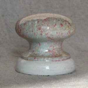 Op de afbeelding: Een keramische knop met een gespikkelde glazuur in tinten wit, groen en rood. De knop heeft een afgeronde bovenkant en een licht uitlopende basis.