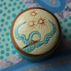 May include: A wooden sphere with a painted design of a blue bird with outstretched wings flying under a cluster of gold stars. The sphere is on a blue and green patterned surface.