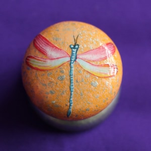 Puede incluir: Una piedra redonda pintada de naranja con una libélula azul pintada sobre ella. La libélula tiene alas rosas y amarillas.