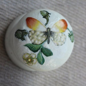 Puede incluir: Dos pomos redondos de madera con un diseño de mariposa pintado. El pomo superior es blanco con una hoja verde y una flor amarilla. El pomo inferior es marrón con una base azul.