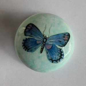 Op de afbeelding: Een ronde, lichtblauwe decoratieve steen met een gedetailleerde blauwe vlinder. De vlinder heeft zwarte en roze accenten en ingewikkelde vleugelpatronen. De steen heeft een glanzende afwerking.