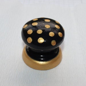 May include: A black wooden cabinet knob with gold polka dots and a gold base. The knob is oval-shaped and sits on a white surface. The knob is a decorative element for furniture.