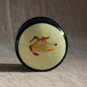 Puede incluir: Un servilletero redondo, negro y amarillo, con una ilustraci&oacute;n de un perro pintado. El perro tiene pelaje naranja, ojos azules y un collar rojo. El servilletero est&aacute; sobre una superficie beige texturizada.