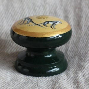 Può includere: Un pomello in legno verde scuro con un bordo dorato e un disegno dipinto di due animali grigi su sfondo giallo.