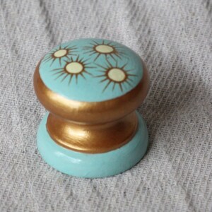 May include: A light blue and gold knob with a decorative design of white sunbursts. The knob is made of wood and has a smooth, rounded surface.