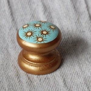 May include: A small, round, gold-painted knob with a blue top. The top is decorated with a pattern of white stars on a light blue background.