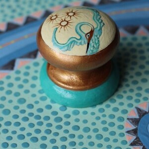 Puede incluir: Un pomo pintado en azul turquesa y dorado con un fondo blanco y un diseño de pájaro azul. El pájaro tiene un cuello largo y un pico largo. Hay tres estrellas amarillas en el pomo.