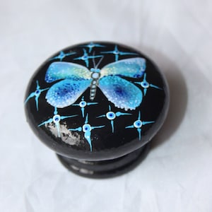 May include: A black wooden knob with a blue butterfly painted on it. The butterfly has white accents and is surrounded by a pattern of white stars on a black background.