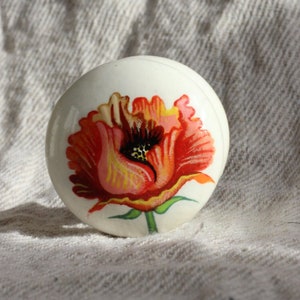 Pode incluir: Puxador de armário em cerâmica branca com um vibrante design de flor de papoula vermelha e laranja. A obra de arte floral inclui folhas verdes e um centro preto, adicionando um toque de decoração inspirada na natureza a qualquer gaveta ou armário.