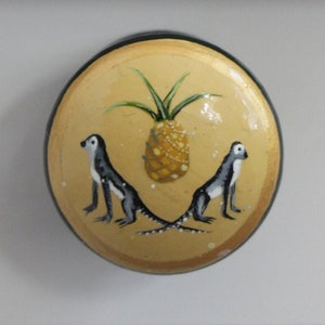 Może przedstawiać: Okrągła, dekoracyjna gałka do szafki z malowanym wzorem. Gałka ma żółte tło z ananasem i dwoma małpami. Małpy są czarno-białe z długimi ogonami. Gałka ma czarną krawędź i złote wykończenie.