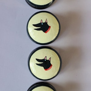 Puede incluir: Cuatro objetos redondos con borde negro y centro amarillo claro. Cada uno presenta una cabeza de perro negra con un collar rojo. La cabeza del perro está estilizada con orejas puntiagudas y un hocico largo. Los objetos están dispuestos verticalmente.