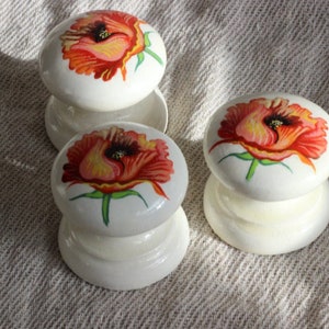 Könnte beinhalten: Drei weiße Keramikknöpfe mit roten Mohnblumen-Designs. Die Knöpfe sind rund und haben einen erhöhten Rand. Die Blumen sind in Rot-, Orange- und Gelbtönen bemalt.