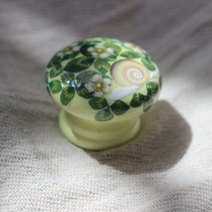 May include: A light green ceramic knob with a painted design of a snail and flowers. The knob has a rounded top and a cylindrical base.