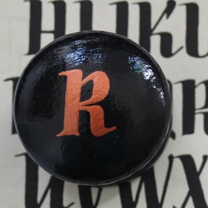 May include: A black, circular knob featuring the letter "R" in orange. The knob is set against a backdrop of black and white lettering. This decorative knob could be used for furniture or as a decorative accent.
