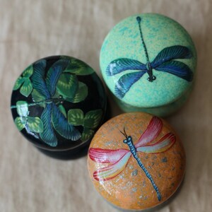 Może przedstawiać: Trzy okrągłe drewniane ozdoby z malowanymi wzorami ważek. Ozdoby są pomalowane w odcieniach zieleni, błękitu i pomarańczy.