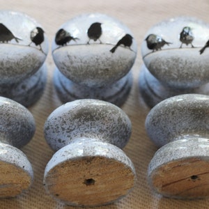 K&ouml;nnte beinhalten: Sechs graue und wei&szlig;e Keramik-Schrankkn&ouml;pfe mit einem Design aus schwarzen V&ouml;geln auf einem Draht. Die Kn&ouml;pfe haben eine Holzbasis.