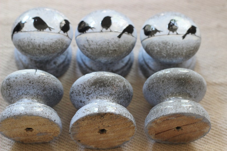 K&ouml;nnte beinhalten: Sechs graue und wei&szlig;e Keramik-Schrankkn&ouml;pfe mit einem Design aus schwarzen V&ouml;geln auf einem Draht. Die Kn&ouml;pfe haben eine Holzbasis.