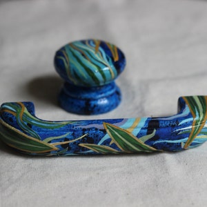 May include: A set of two blue painted cabinet knobs with a green and gold leaf design. The knobs are made of wood and have a glossy finish.