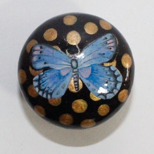 Pode incluir: Uma pedra redonda, pintada de preto com bolinhas douradas e uma borboleta azul pintada no topo.