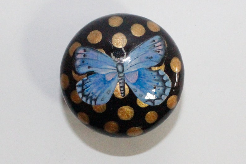 Pode incluir: Uma pedra redonda, pintada de preto com bolinhas douradas e uma borboleta azul pintada no topo.