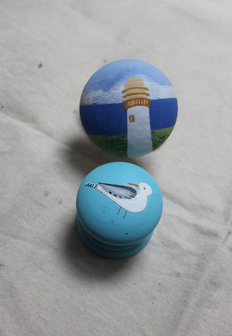 May include: Two hand-painted wooden knobs. One knob features a lighthouse scene with a blue sky and sea. The other knob is light blue with a painted seagull. These decorative knobs are perfect for cabinets or drawers.