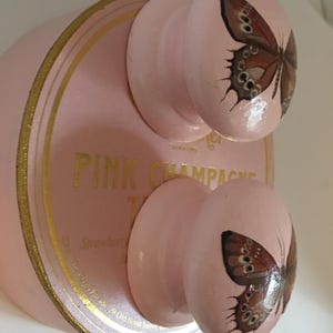 Peut inclure: Bouchon de bouteille de champagne rose avec deux boutons, chacun avec un motif de papillon peint. Le bouchon a des lettres et des bordures dor&eacute;es. Le texte sur le bouchon indique "PINK CHAMPAGNE".
