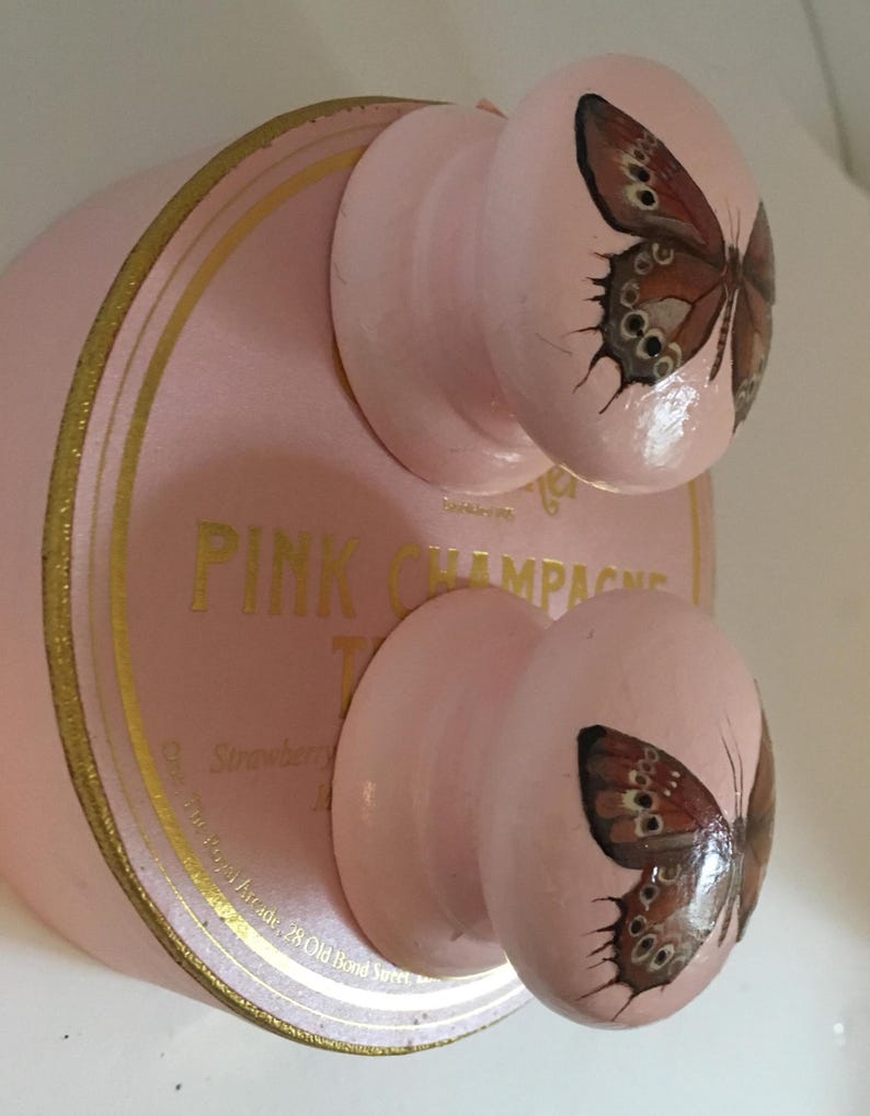 Peut inclure: Bouchon de bouteille de champagne rose avec deux boutons, chacun avec un motif de papillon peint. Le bouchon a des lettres et des bordures dor&eacute;es. Le texte sur le bouchon indique "PINK CHAMPAGNE".