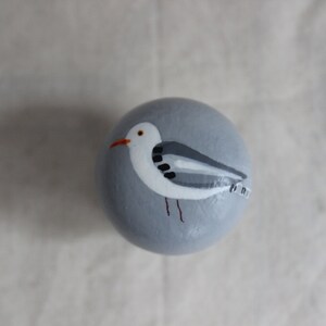K&ouml;nnte beinhalten: Ein handbemalter grauer Stein mit einer darauf gemalten wei&szlig;en M&ouml;we. Die M&ouml;we hat schwarze Augen und einen roten Schnabel.