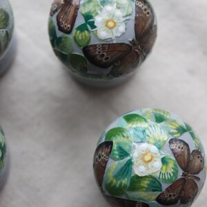 Puede incluir: Tres adornos redondos con fondo azul y una flor blanca con centro amarillo. Cada adorno est&aacute; decorado con mariposas marrones y hojas verdes.