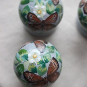 Puede incluir: Tres adornos redondos con un fondo gris claro y dise&ntilde;os pintados de mariposas y flores blancas con hojas verdes.