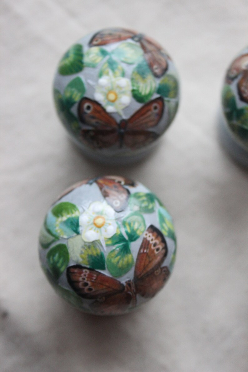 Puede incluir: Tres adornos redondos con un fondo gris claro y dise&ntilde;os pintados de mariposas y flores blancas con hojas verdes.