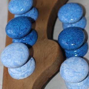Puede incluir: Una tabla de servir de madera con dos conjuntos de piedras decorativas ovales azules moteadas. Las piedras varían en tonos de azul, de claro a oscuro, y están dispuestas en pilas verticales. La madera tiene un color marrón claro natural.